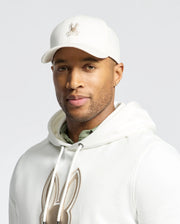 A man poses against a plain background, exuding quiet luxury in a white hoodie with a bunny design and the Psycho Bunny MENS NEW CASTLE GRAPHIC BASEBALL CAP (B6A547F200), showcasing a similar logo.