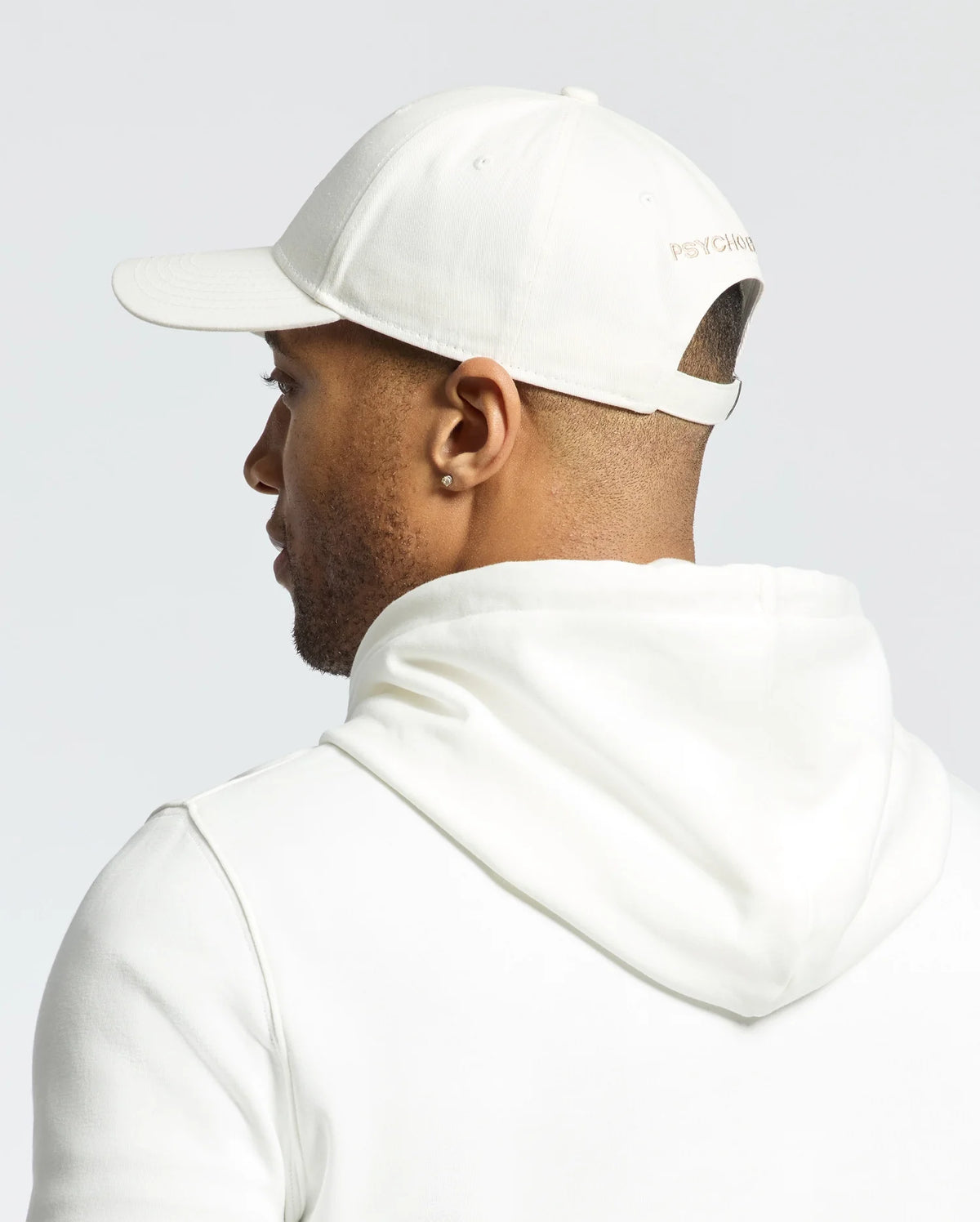 A person is shown from the back wearing a white hooded sweatshirt and the Psycho Bunny MENS NEW CASTLE GRAPHIC BASEBALL CAP, made of cotton twill with an adjustable strap and subtle side text, embodying quiet luxury against a plain, light-colored background.