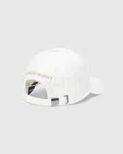 The Men's New Castle Graphic Baseball Cap by Psycho Bunny features white cotton twill, an adjustable strap with a metal buckle at the back, "PSYCHO BUNNY" embroidered in pink on the back panel for a luxurious touch, and a small logo tag near the buckle.