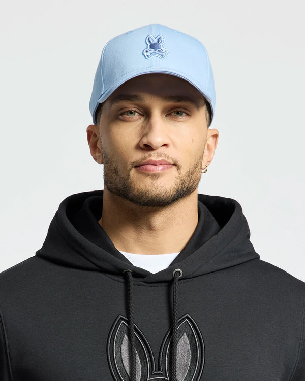 A person in a cotton twill light blue MENS NEW CASTLE GRAPHIC BASEBALL CAP by Psycho Bunny, embroidered with a bunny logo, and wearing a black hoodie with a similar design, looks forward against a plain light background that exudes quiet luxury.