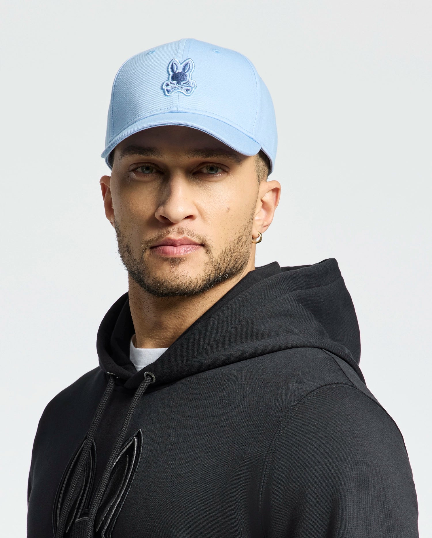 A man exuding quiet luxury wears the MENS NEW CASTLE GRAPHIC BASEBALL CAP by Psycho Bunny in light blue, paired with a black hoodie and small hoop earrings. Against a white background, his neutral expression enhances his understated style.
