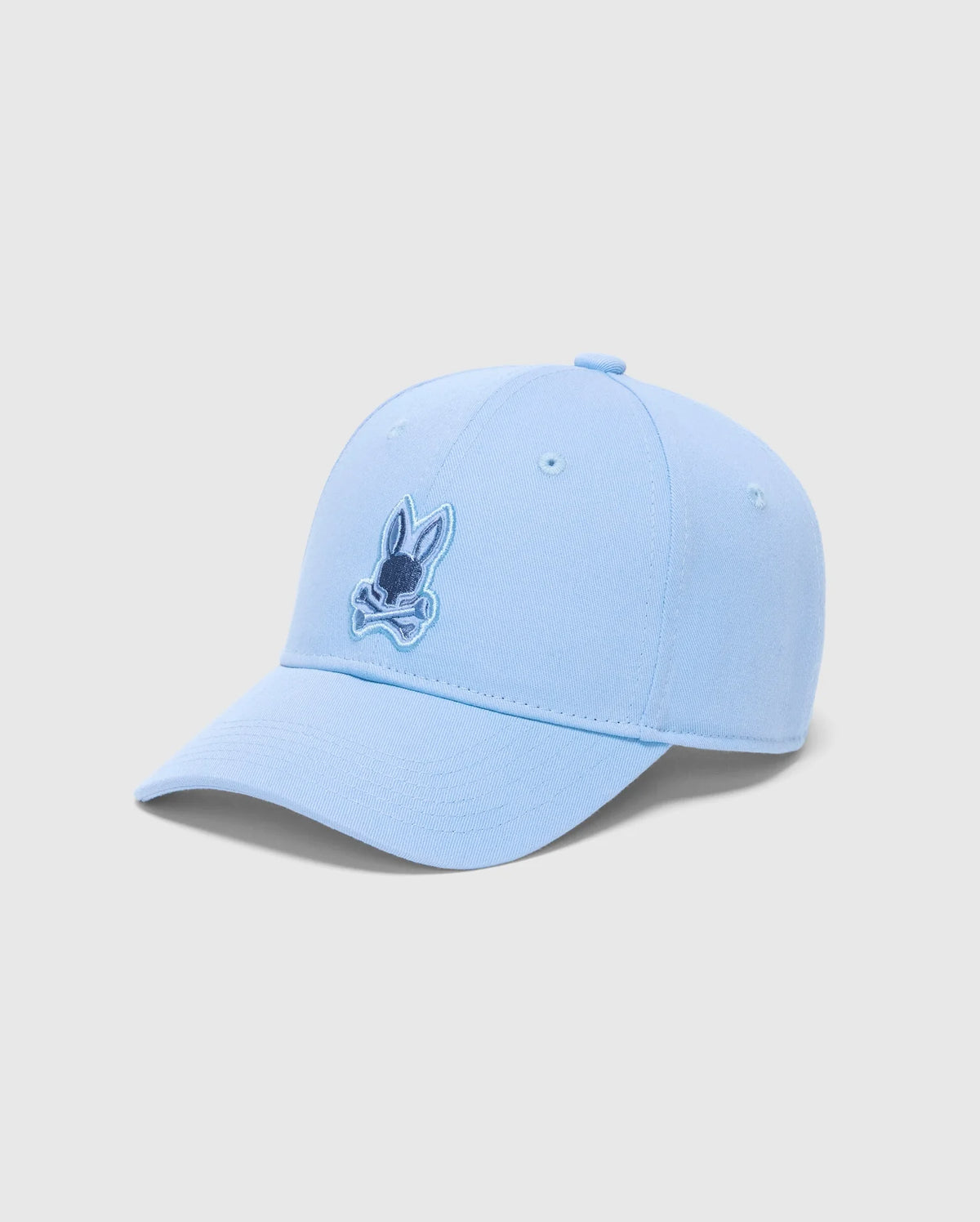 The MENS NEW CASTLE GRAPHIC BASEBALL CAP (B6A547F200) by Psycho Bunny is a light blue cotton twill cap with a rabbit's head and crossbones in darker blue on the front. Set against a plain white backdrop, it captures quiet luxury through its subtle yet striking design.