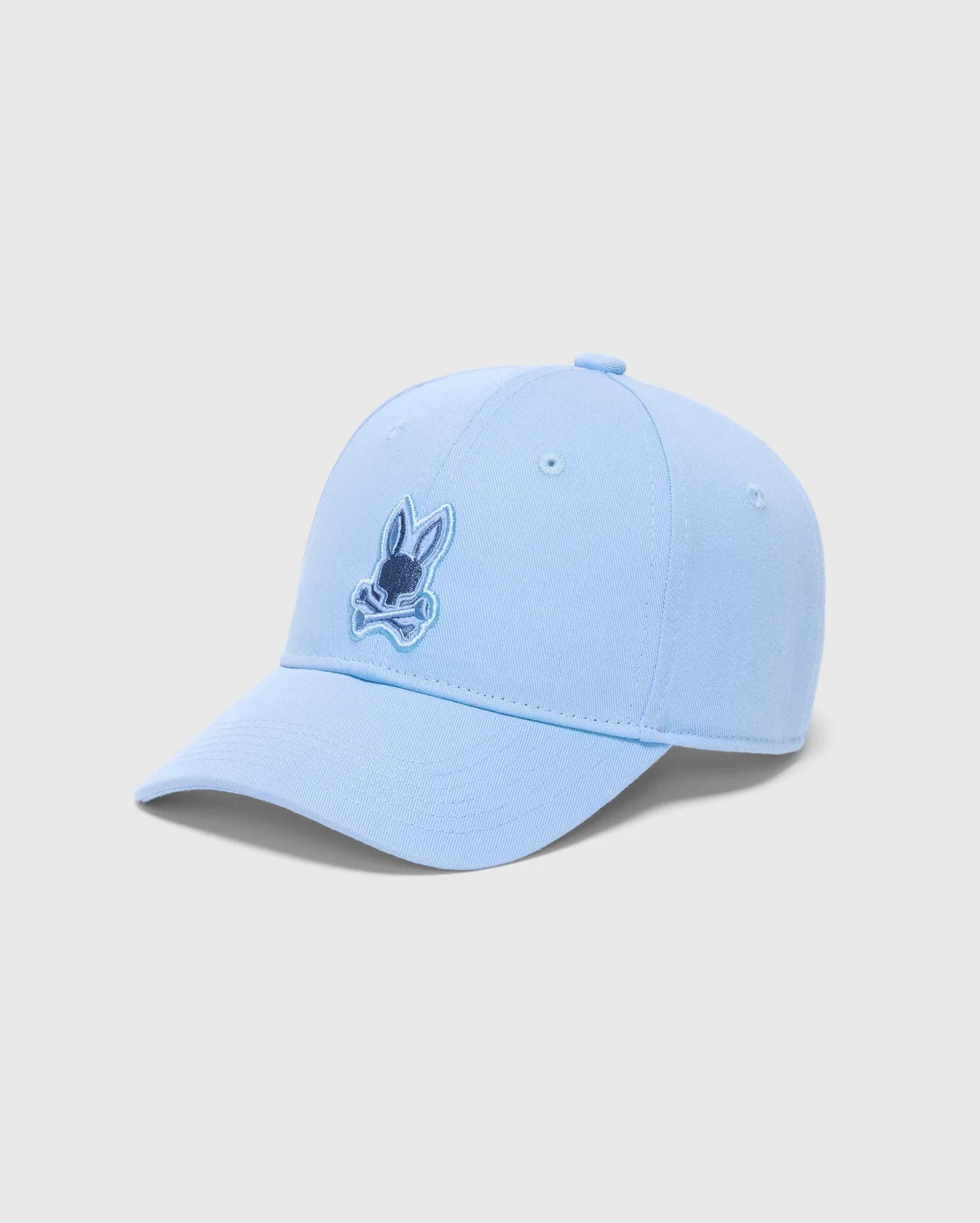 The MENS NEW CASTLE GRAPHIC BASEBALL CAP (B6A547F200) by Psycho Bunny is a light blue cotton twill cap with a rabbit's head and crossbones in darker blue on the front. Set against a plain white backdrop, it captures quiet luxury through its subtle yet striking design.
