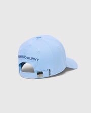 The MENS NEW CASTLE GRAPHIC BASEBALL CAP - B6A547F200 by Psycho Bunny embodies quiet luxury with PSYCHO BUNNY embroidered on the light blue cotton twill and features an adjustable strap with a metal clasp, showcasing an engraved logo against a plain gray background.