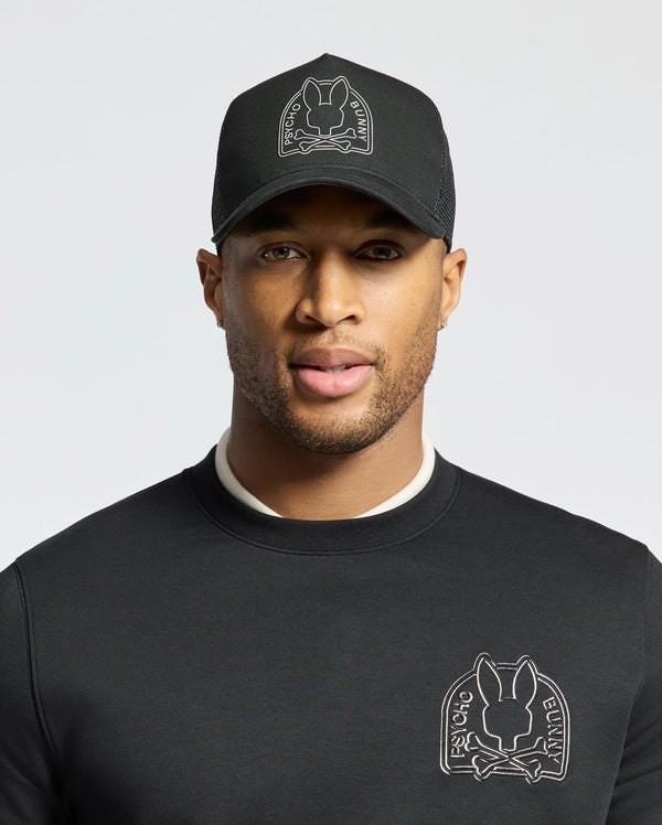 A person wearing a black sweatshirt and a Psycho Bunny MENS MELBOURNE GRAPHIC TRUCKER HAT, both featuring the brand's embroidered bunny logo. The hat's snap-back strap provides a casual touch against the plain, light-colored backdrop.