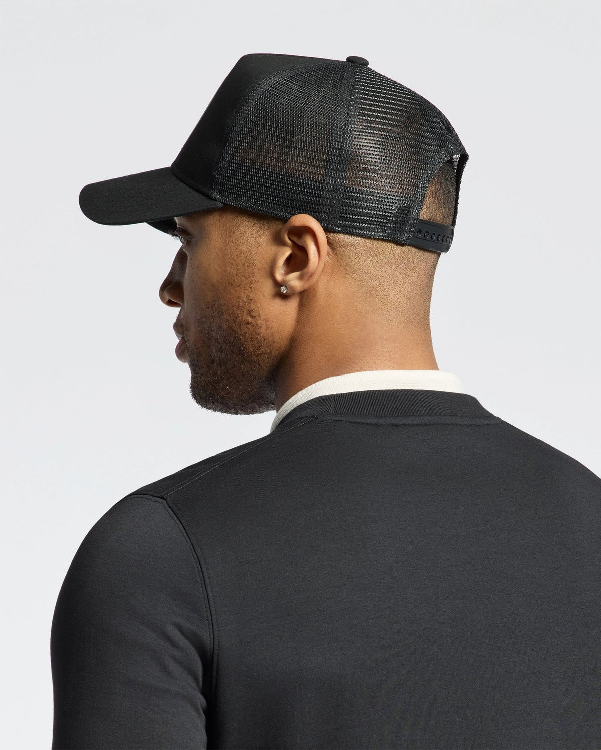 A man in a dark shirt faces away, wearing a Psycho Bunny MENS MELBOURNE GRAPHIC TRUCKER HAT with a snap-back strap.