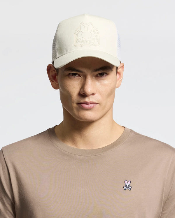 A person wears a beige Psycho Bunny MENS MELBOURNE GRAPHIC TRUCKER HAT with an embossed logo and snap-back strap, paired with a tan shirt featuring a small embroidered bunny insignia on the chest, against a plain, light-colored background.