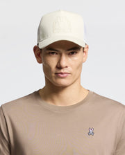 A person wears a beige Psycho Bunny MENS MELBOURNE GRAPHIC TRUCKER HAT with an embossed logo and snap-back strap, paired with a tan shirt featuring a small embroidered bunny insignia on the chest, against a plain, light-colored background.