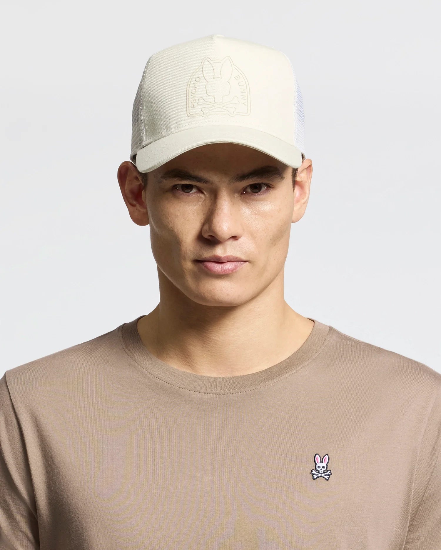 A person wears a beige Psycho Bunny MENS MELBOURNE GRAPHIC TRUCKER HAT with an embossed logo and snap-back strap, paired with a tan shirt featuring a small embroidered bunny insignia on the chest, against a plain, light-colored background.
