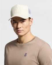 A man sports a beige Psycho Bunny MENS MELBOURNE GRAPHIC TRUCKER HAT with a snap-back strap and embossed logo, paired with a beige long-sleeve shirt featuring an embroidered bunny icon above its cuff. The light gray backdrop highlights the understated style.