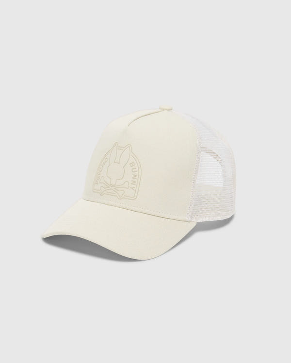 The MENS MELBOURNE GRAPHIC TRUCKER HAT by Psycho Bunny is light beige with a mesh back and curved brim. It features a subtle embossed bunny and crossbones on the front, complemented by an adjustable snap-back strap for the perfect fit.