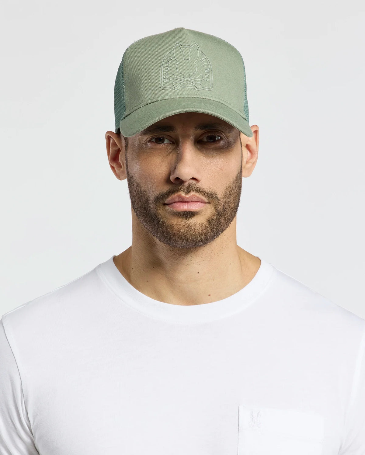 A bearded man dons a Psycho Bunny MENS MELBOURNE GRAPHIC TRUCKER HAT - B6A548F200 in light green with an embroidered icon, teamed with a plain white t-shirt for a stylish look.