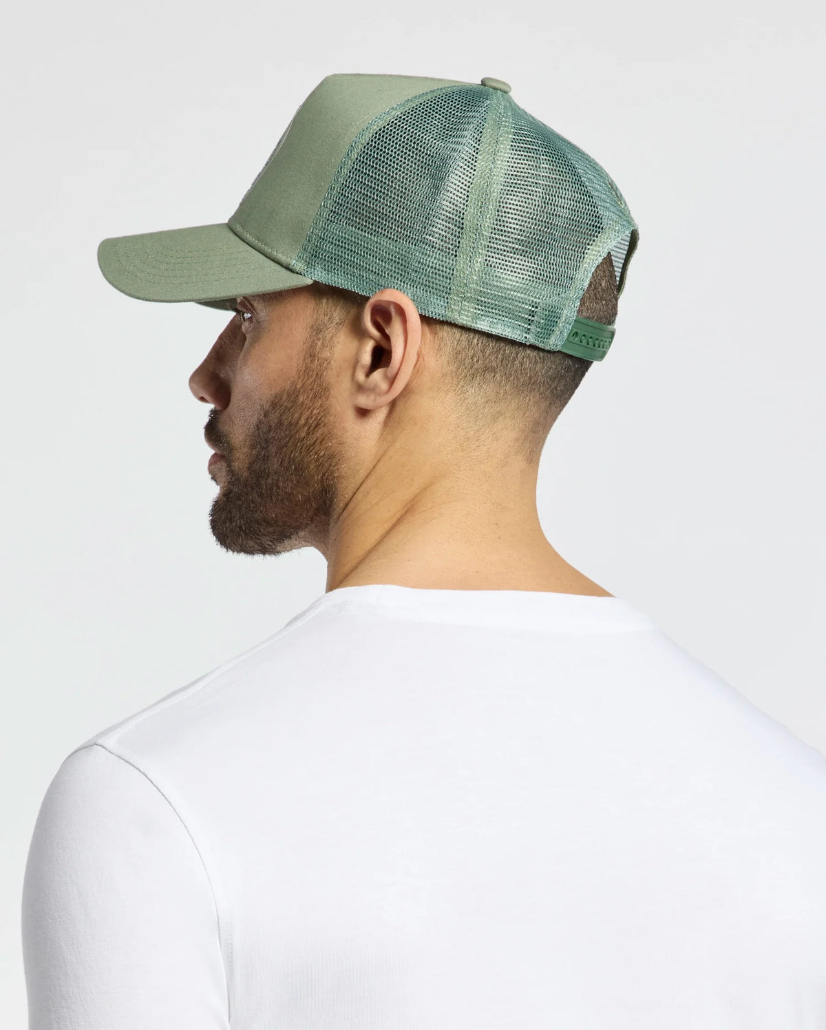 A person wearing a Psycho Bunny light green trucker hat (MENS MELBOURNE GRAPHIC, B6A548F200) and a white shirt is seen from behind against a plain gray background.