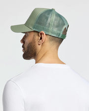 A person wearing a Psycho Bunny light green trucker hat (MENS MELBOURNE GRAPHIC, B6A548F200) and a white shirt is seen from behind against a plain gray background.