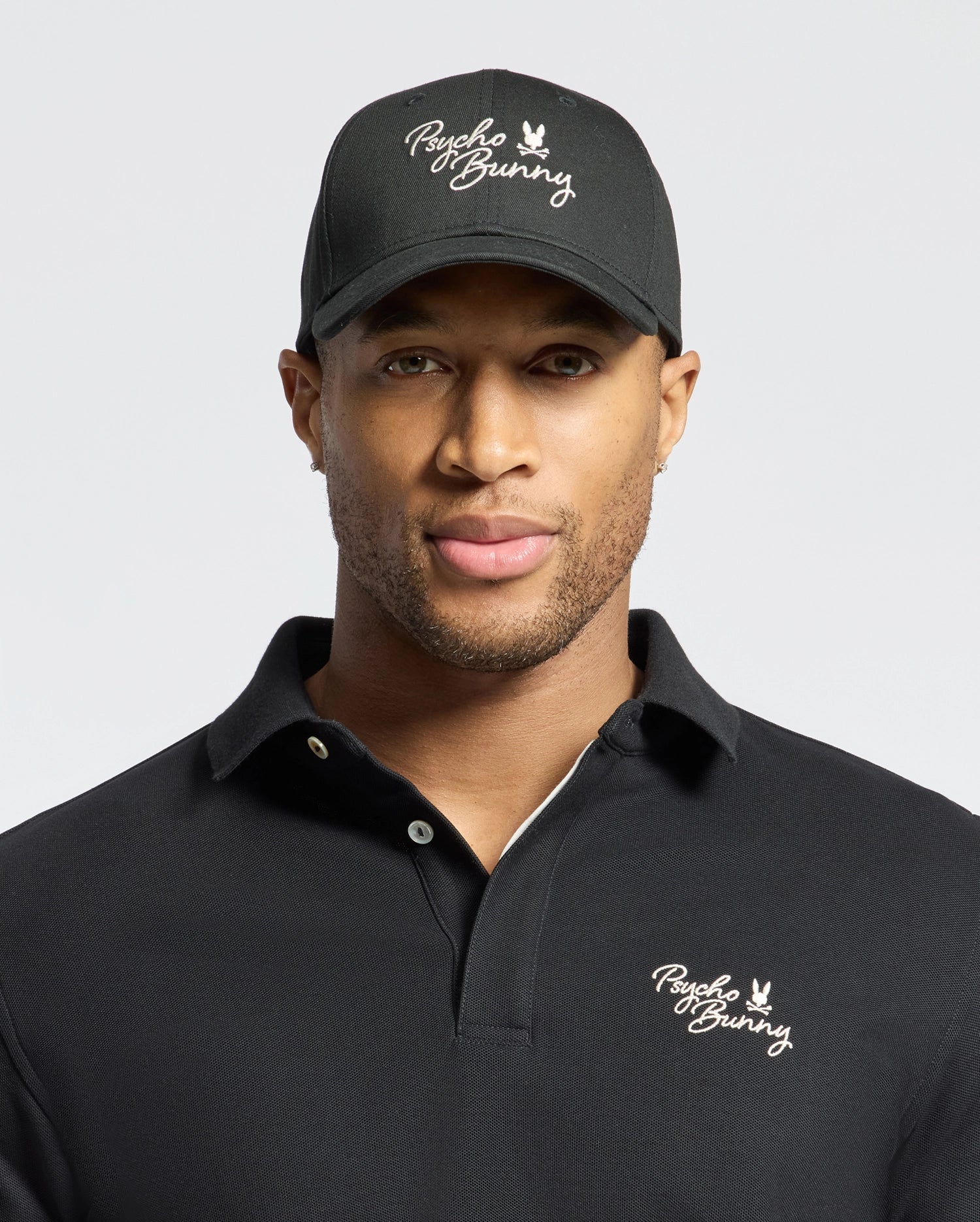 A man wears a Psycho Bunny black 100% cotton polo shirt and the MENS DESERT BASEBALL CAP (B6A549F200) with the logo, set against a plain backdrop.
