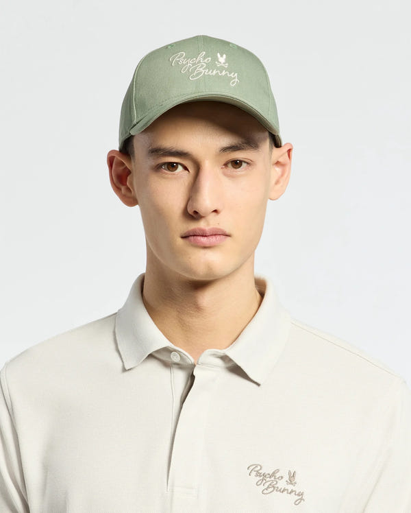 Wearing a Psycho Bunny MENS DESERT BASEBALL CAP in sage green with an embroidered wordmark and adjustable strap, paired with a light beige polo shirt featuring the same logo, they stand against a plain light background.