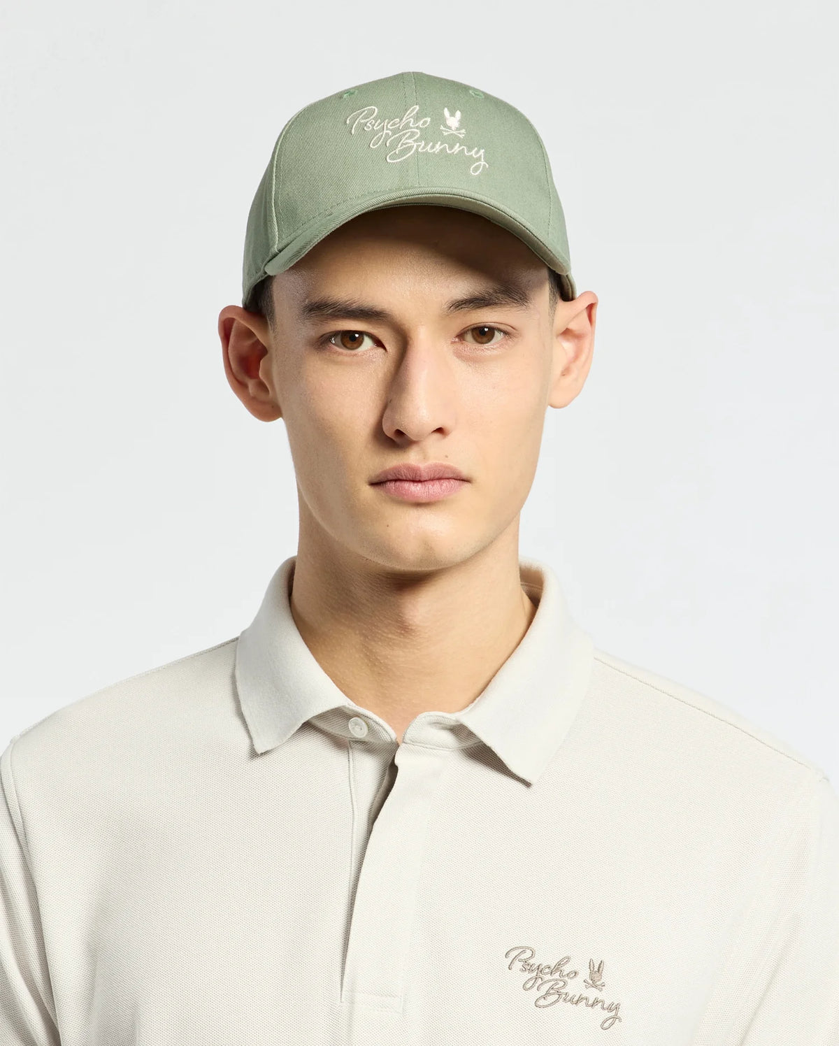 Wearing a Psycho Bunny MENS DESERT BASEBALL CAP in sage green with an embroidered wordmark and adjustable strap, paired with a light beige polo shirt featuring the same logo, they stand against a plain light background.