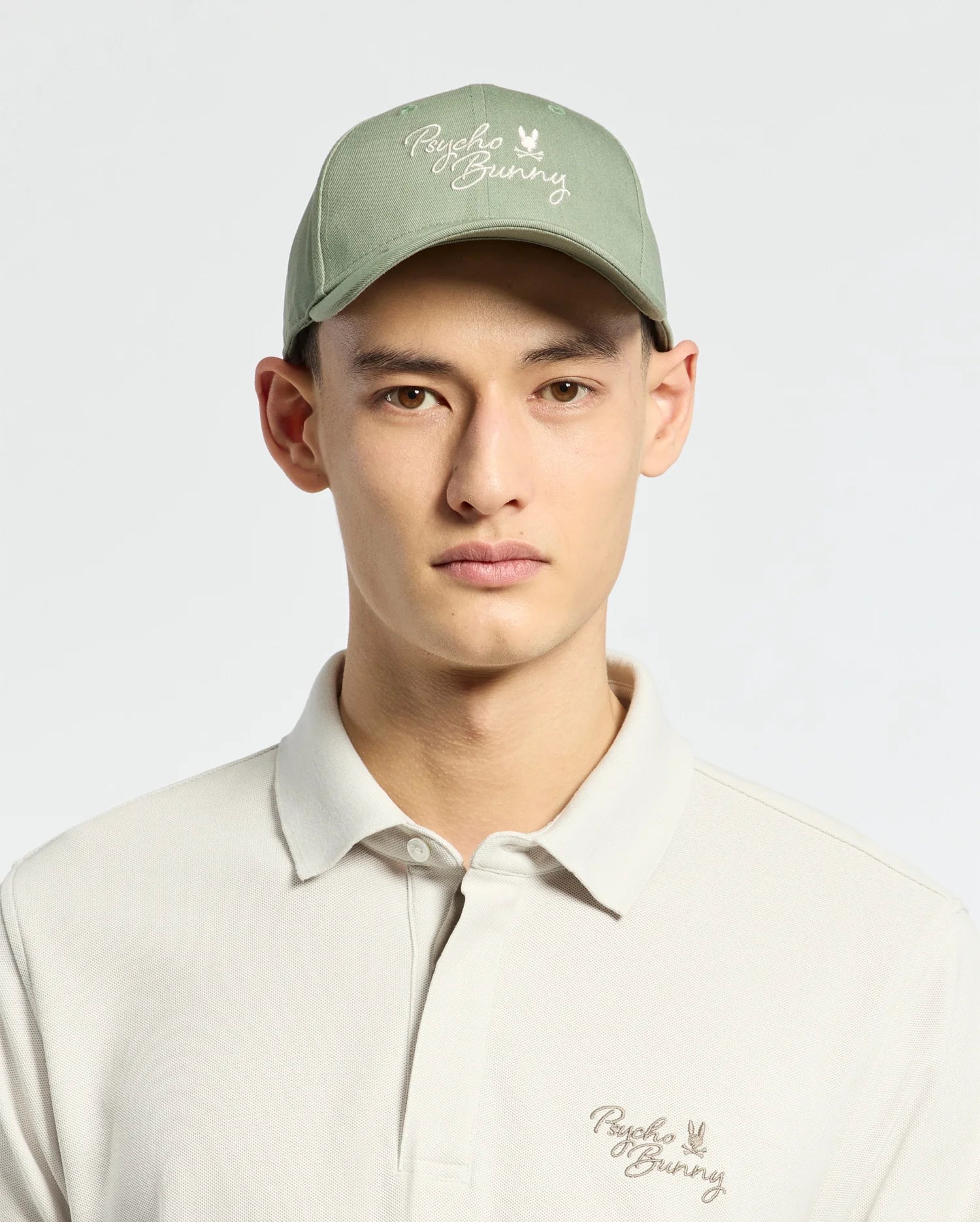 Wearing a Psycho Bunny MENS DESERT BASEBALL CAP in sage green with an embroidered wordmark and adjustable strap, paired with a light beige polo shirt featuring the same logo, they stand against a plain light background.