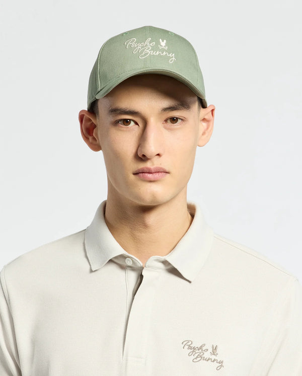 Wearing a Psycho Bunny MENS DESERT BASEBALL CAP in sage green with an embroidered wordmark and adjustable strap, paired with a light beige polo shirt featuring the same logo, they stand against a plain light background.