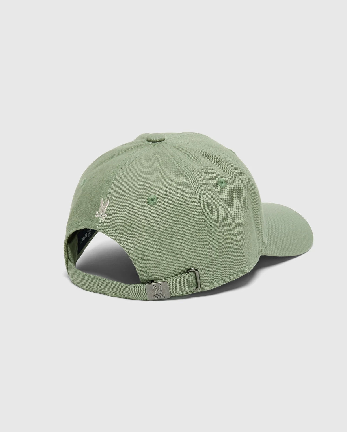The Psycho Bunny MENS DESERT BASEBALL CAP - B6A549F200 is shown from the back, displaying a light green cotton material with an adjustable metal buckle strap. A small embroidered wordmark appears above the opening, all set against a plain gray background.
