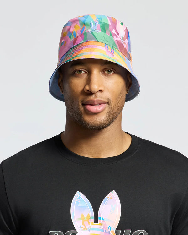 A person sports a Psycho Bunny MENS MILOS BUCKET REVERSIBLE HAT (B6A550F200) with vibrant geometric prints and a black shirt featuring large bunny ears against a plain light grey background.