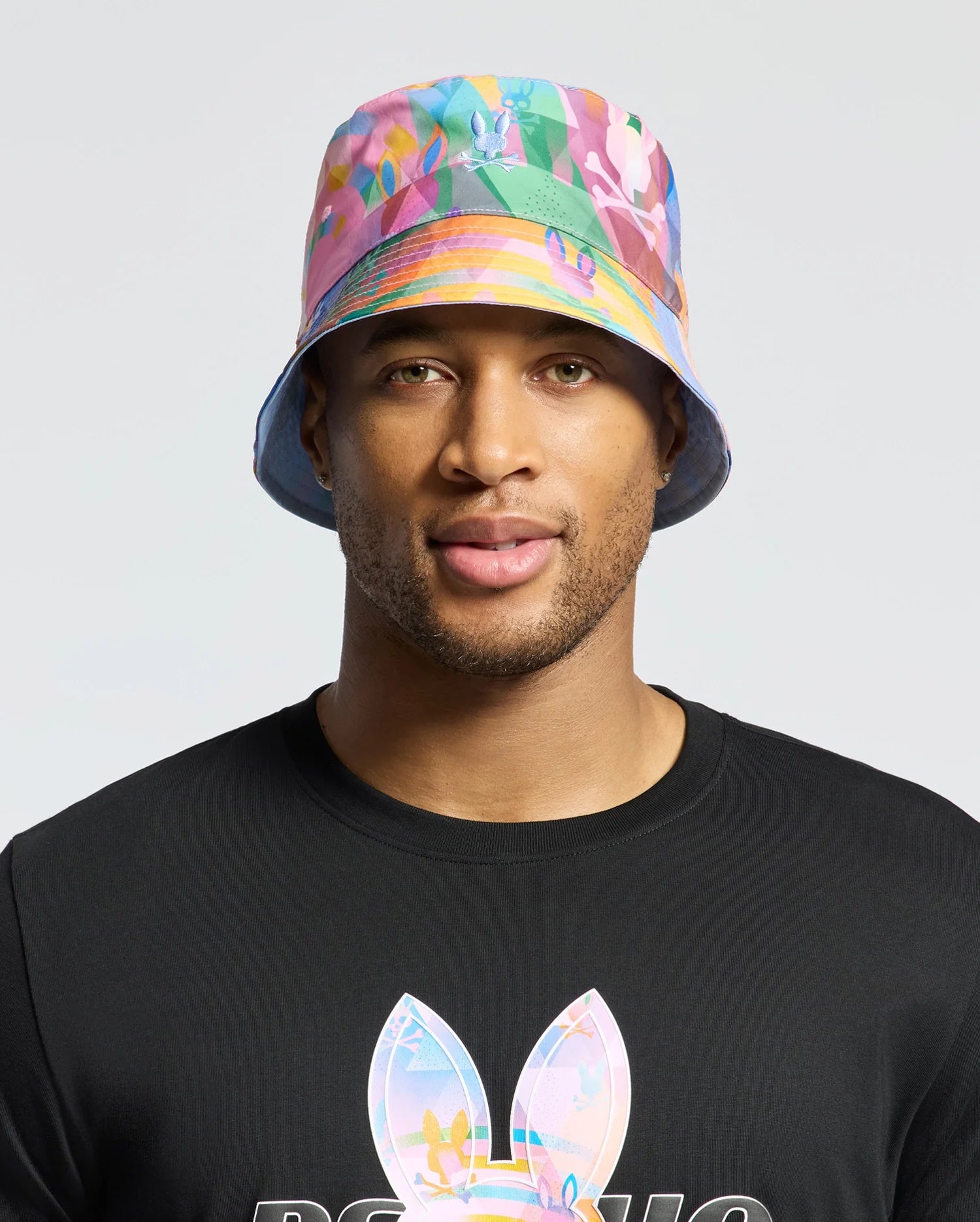 A person sports a Psycho Bunny MENS MILOS BUCKET REVERSIBLE HAT (B6A550F200) with vibrant geometric prints and a black shirt featuring large bunny ears against a plain light grey background.