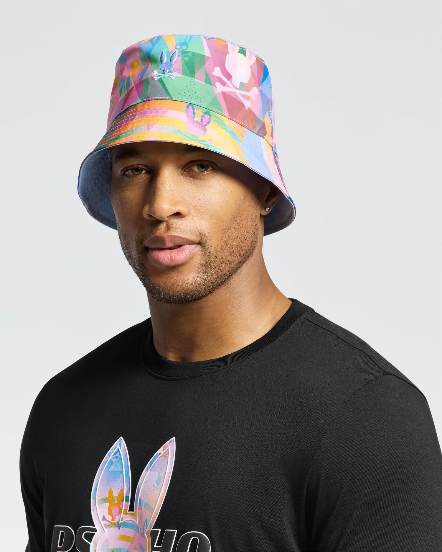 A person stands against a plain background wearing a vibrant Psycho Bunny MENS MILOS BUCKET REVERSIBLE HAT - B6A550F200 and a black t-shirt with a geometric print.