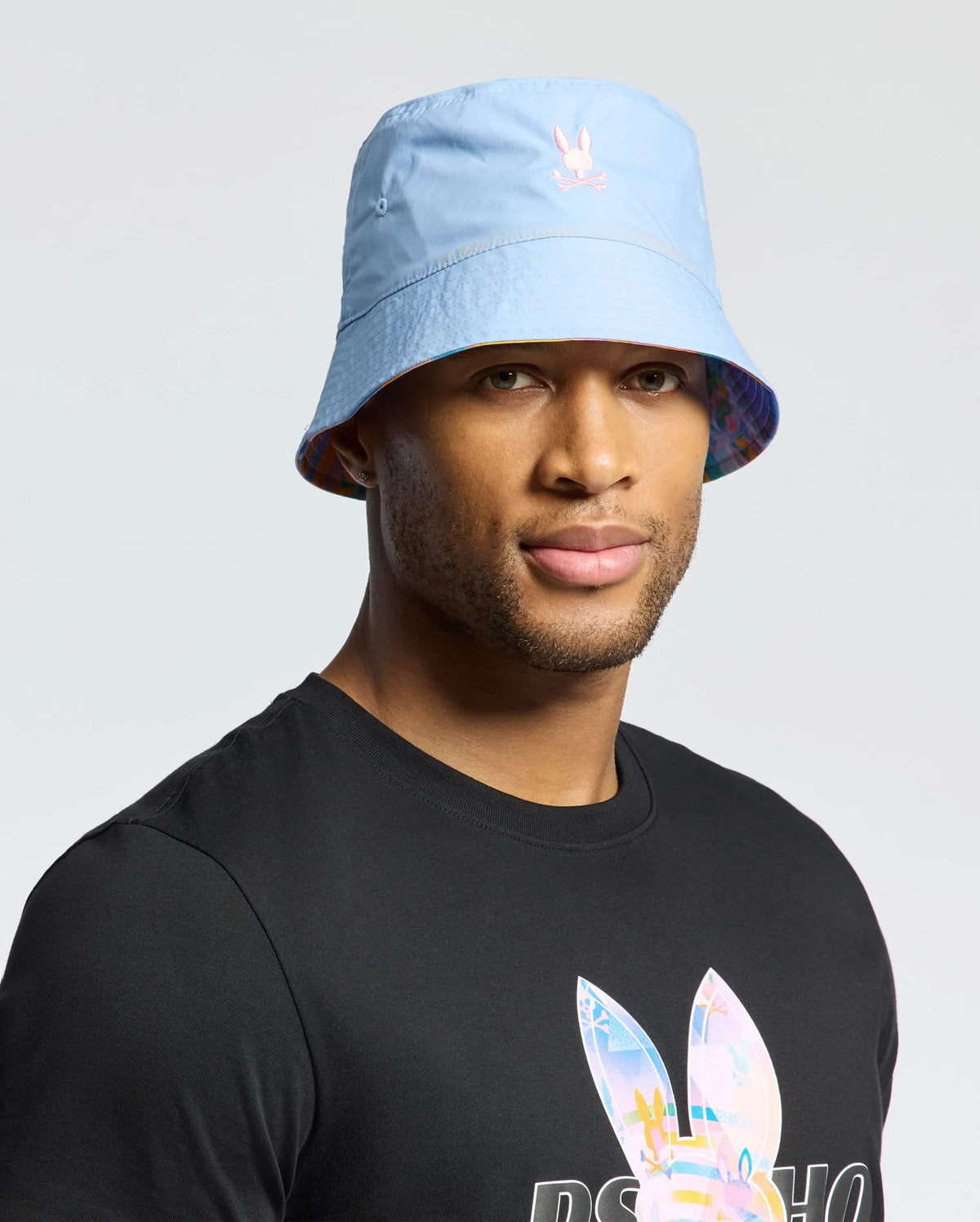 A person wears a light blue Psycho Bunny Milos bucket hat with a small embroidered logo and a black T-shirt featuring a colorful geometric print. The backdrop is plain light gray.