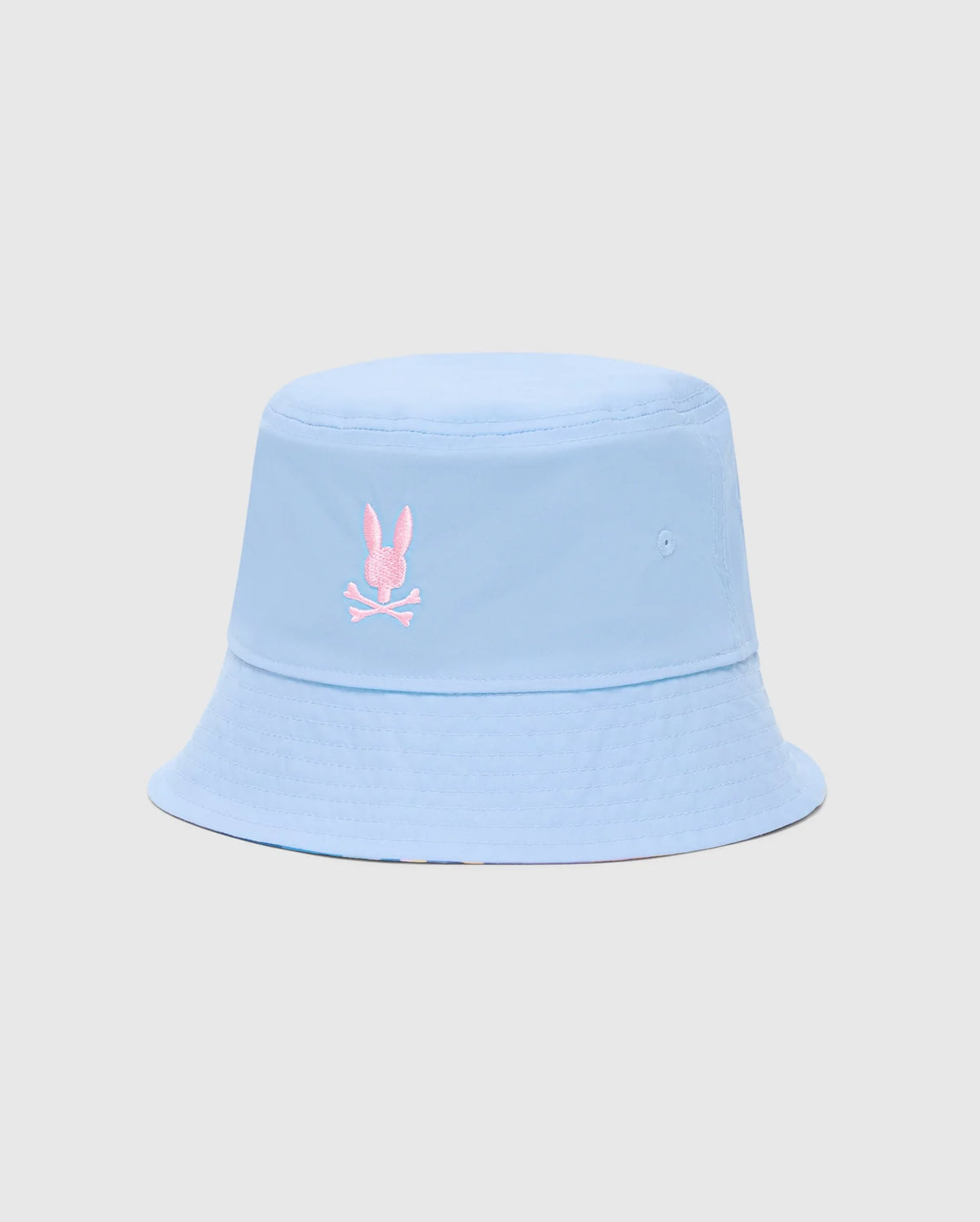 The Psycho Bunny MENS MILOS BUCKET REVERSIBLE HAT (B6A550F200) features light blue fabric with a pink embroidered stylized rabbit above crossed bones on the front. It has a subtle texture, visible stitching along the brim, and is set against a plain gray background.