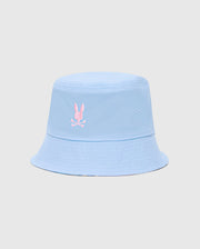 The Psycho Bunny MENS MILOS BUCKET REVERSIBLE HAT (B6A550F200) features light blue fabric with a pink embroidered stylized rabbit above crossed bones on the front. It has a subtle texture, visible stitching along the brim, and is set against a plain gray background.