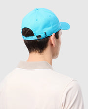 A person wearing the Psycho Bunny MENS LOUISE BASEBALL CAP - B6A551C200, a light blue cotton twill baseball cap with an embroidered logo and an adjustable strap featuring a black buckle, along with a light-colored polo shirt that has a beige collar, faces away. The background is plain light gray.