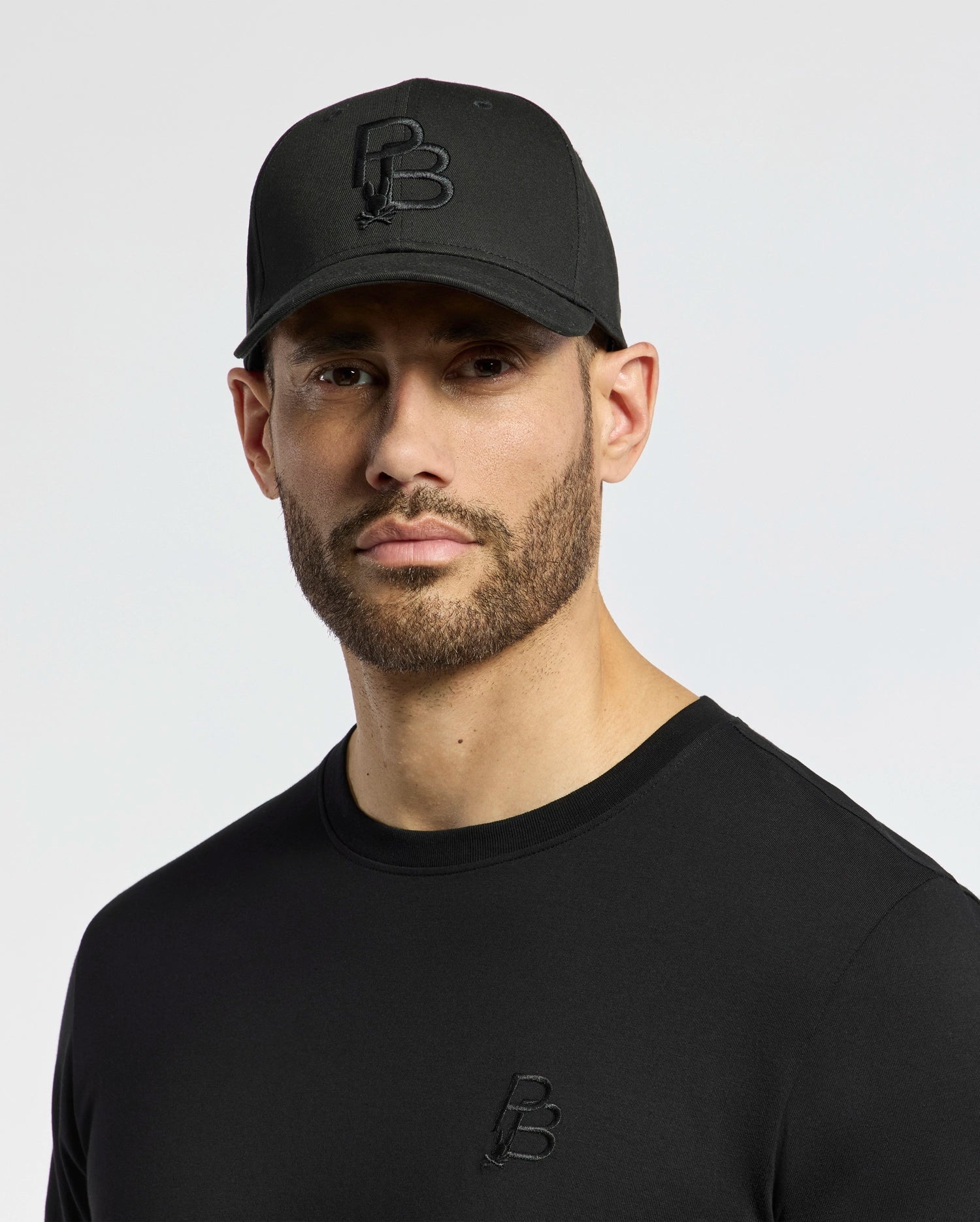 A bearded person wears a Psycho Bunny black cotton twill cap with adjustable backstrap and a black shirt, both showcasing the PB-embroidered design. The background is plain and light-colored.