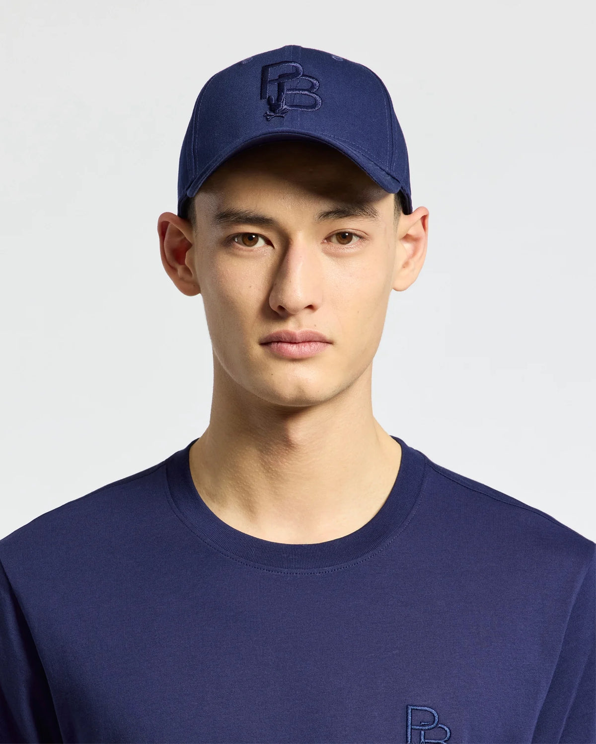 A person wears the MENS NICHOLSON PB BUNNY GRAPHIC BASEBALL CAP by Psycho Bunny, featuring a blue cotton twill construction with an adjustable backstrap, along with a matching blue shirt set against a light background.