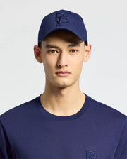 A person wears the MENS NICHOLSON PB BUNNY GRAPHIC BASEBALL CAP by Psycho Bunny, featuring a blue cotton twill construction with an adjustable backstrap, along with a matching blue shirt set against a light background.