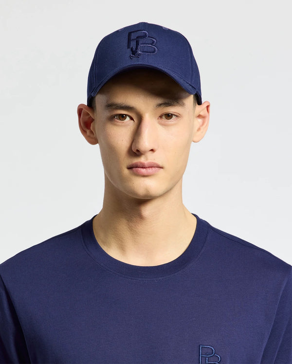 A person wears the MENS NICHOLSON PB BUNNY GRAPHIC BASEBALL CAP by Psycho Bunny, featuring a blue cotton twill construction with an adjustable backstrap, along with a matching blue shirt set against a light background.