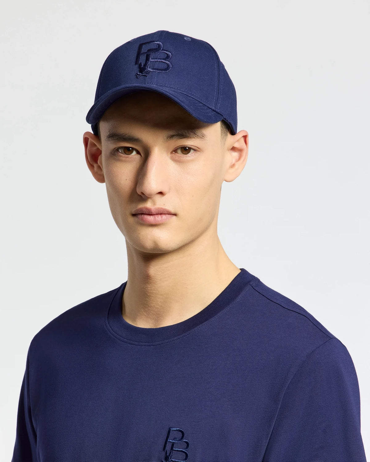 A person sports a Psycho Bunny MENS NICHOLSON PB BUNNY GRAPHIC BASEBALL CAP - B6A553F200, a blue cotton twill cap with embroidery, paired with a matching shirt. The cap has an adjustable backstrap for comfort against the plain, light background.