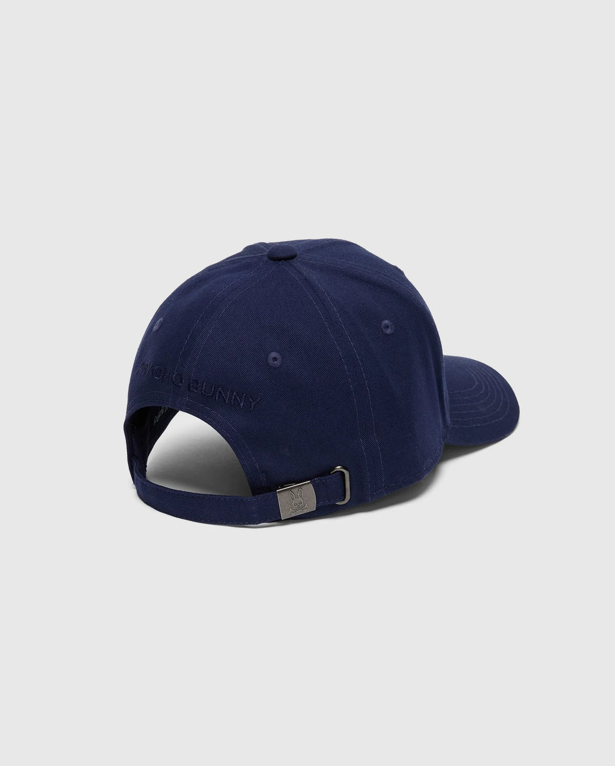 The Psycho Bunny MENS NICHOLSON PB BUNNY GRAPHIC BASEBALL CAP - B6A553F200 is a navy blue cotton twill cap featuring a subtle embroidered design. Viewed from the back, it reveals an adjustable strap with a metal buckle, adding sophistication to its classic look.