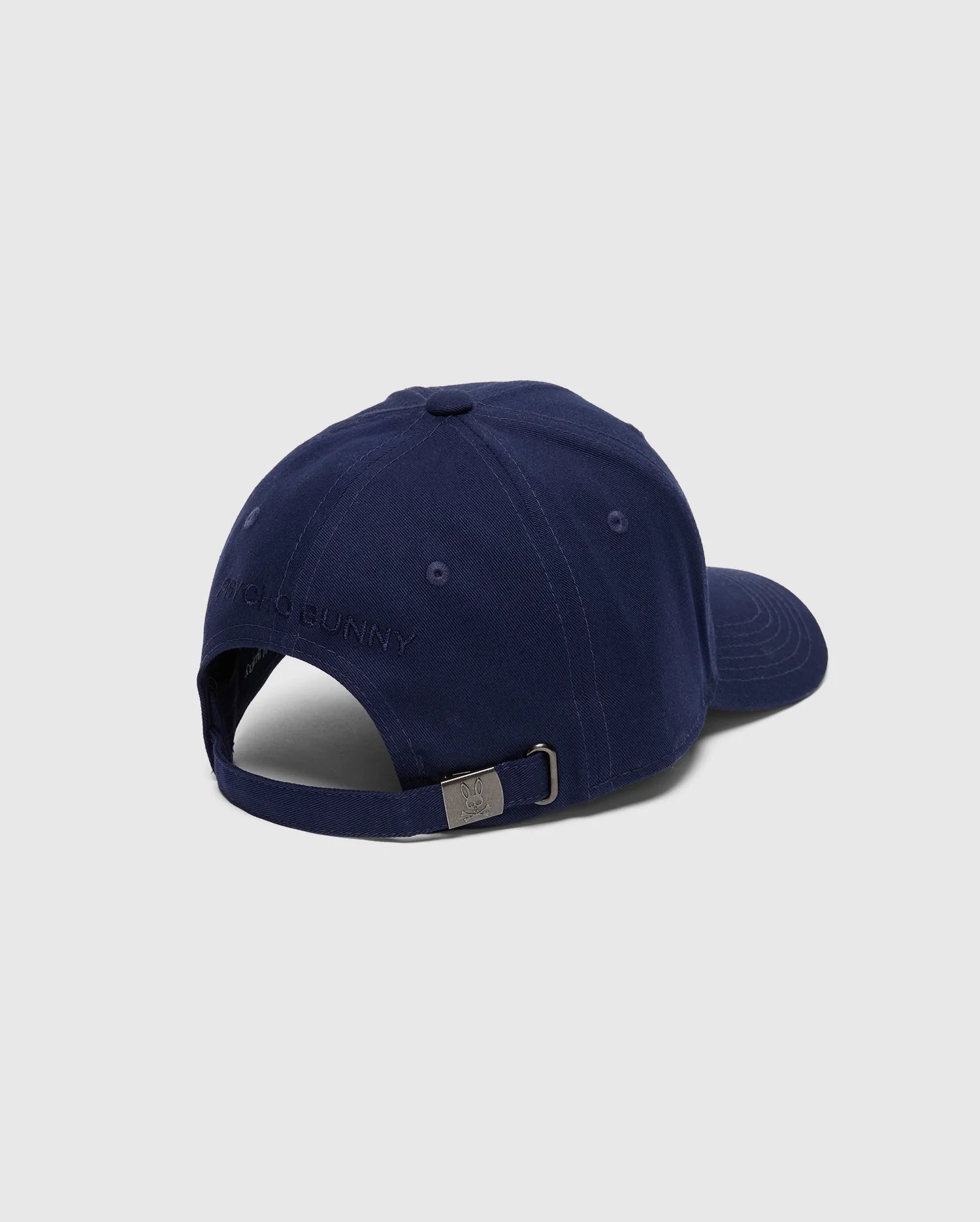 The Psycho Bunny MENS NICHOLSON PB BUNNY GRAPHIC BASEBALL CAP - B6A553F200 is a navy blue cotton twill cap featuring a subtle embroidered design. Viewed from the back, it reveals an adjustable strap with a metal buckle, adding sophistication to its classic look.
