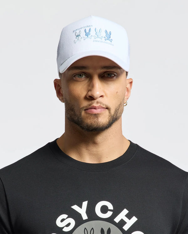 A bearded man sports a black cartoon character tee and a Psycho Bunny MENS OCEANIA EMBROIDERED GRAPHIC TRUCKER HAT with a bunny graphic, complemented by a hoop earring. He exudes street style against a plain backdrop.