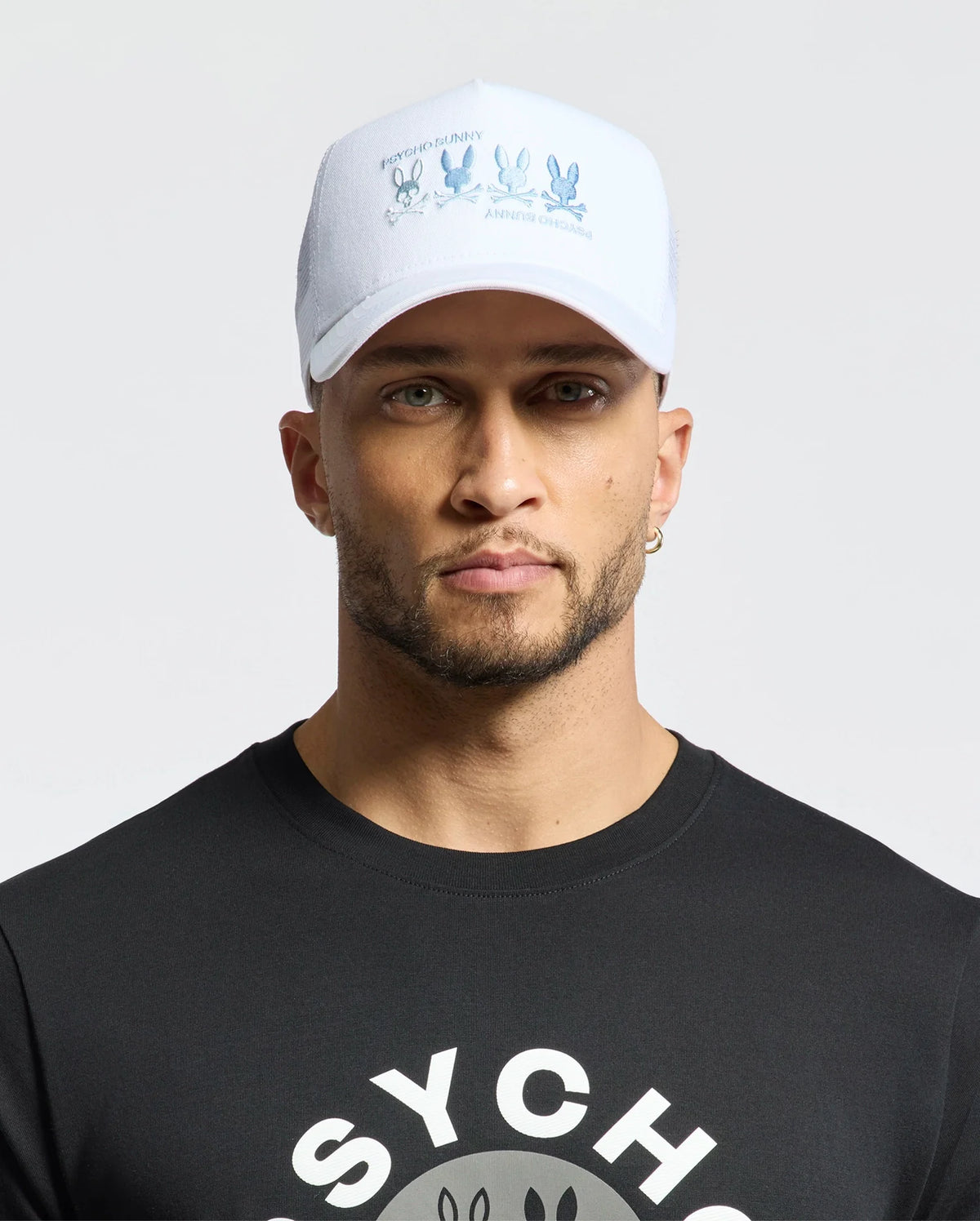 A bearded man sports a black cartoon character tee and a Psycho Bunny MENS OCEANIA EMBROIDERED GRAPHIC TRUCKER HAT with a bunny graphic, complemented by a hoop earring. He exudes street style against a plain backdrop.