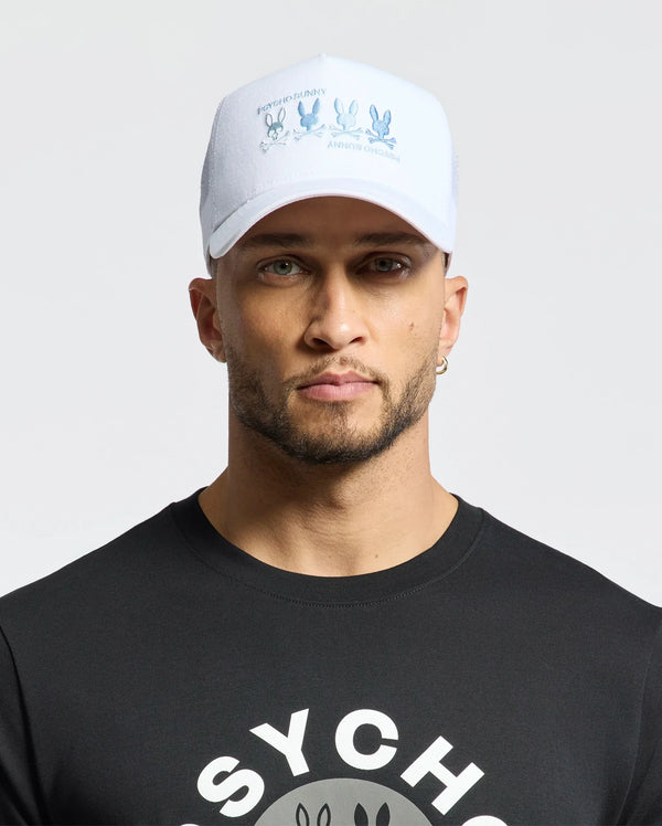 A bearded man sports a black cartoon character tee and a Psycho Bunny MENS OCEANIA EMBROIDERED GRAPHIC TRUCKER HAT with a bunny graphic, complemented by a hoop earring. He exudes street style against a plain backdrop.
