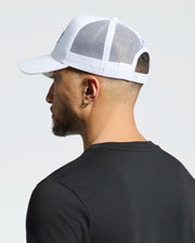 A person in a black t-shirt faces away from the camera, wearing a Psycho Bunny Mens Oceania Embroidered Graphic Trucker Hat (B6A554F200). The white mesh baseball cap enhances the streetwear vibe against the light backdrop.