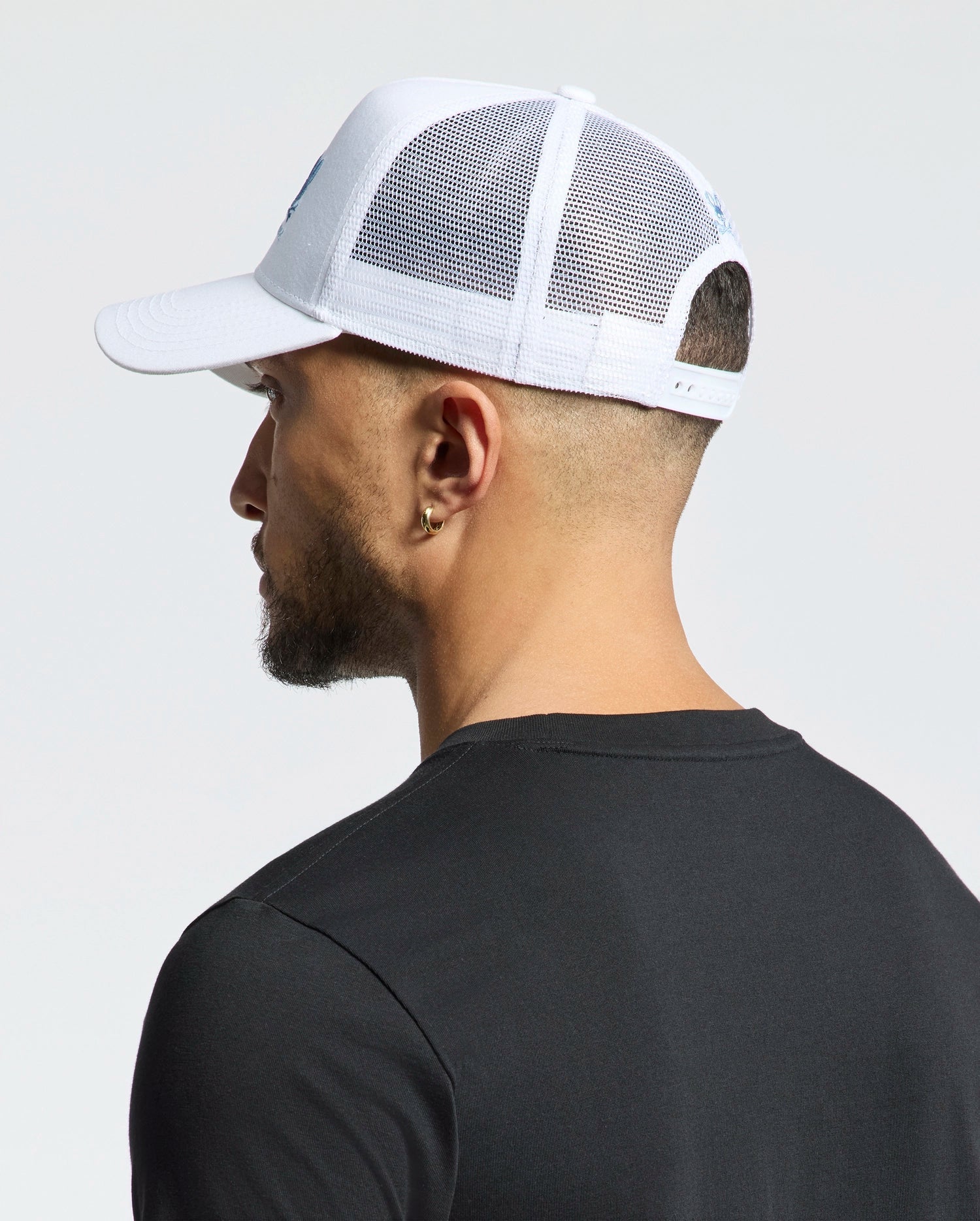 A person in a black t-shirt faces away from the camera, wearing a Psycho Bunny Mens Oceania Embroidered Graphic Trucker Hat (B6A554F200). The white mesh baseball cap enhances the streetwear vibe against the light backdrop.