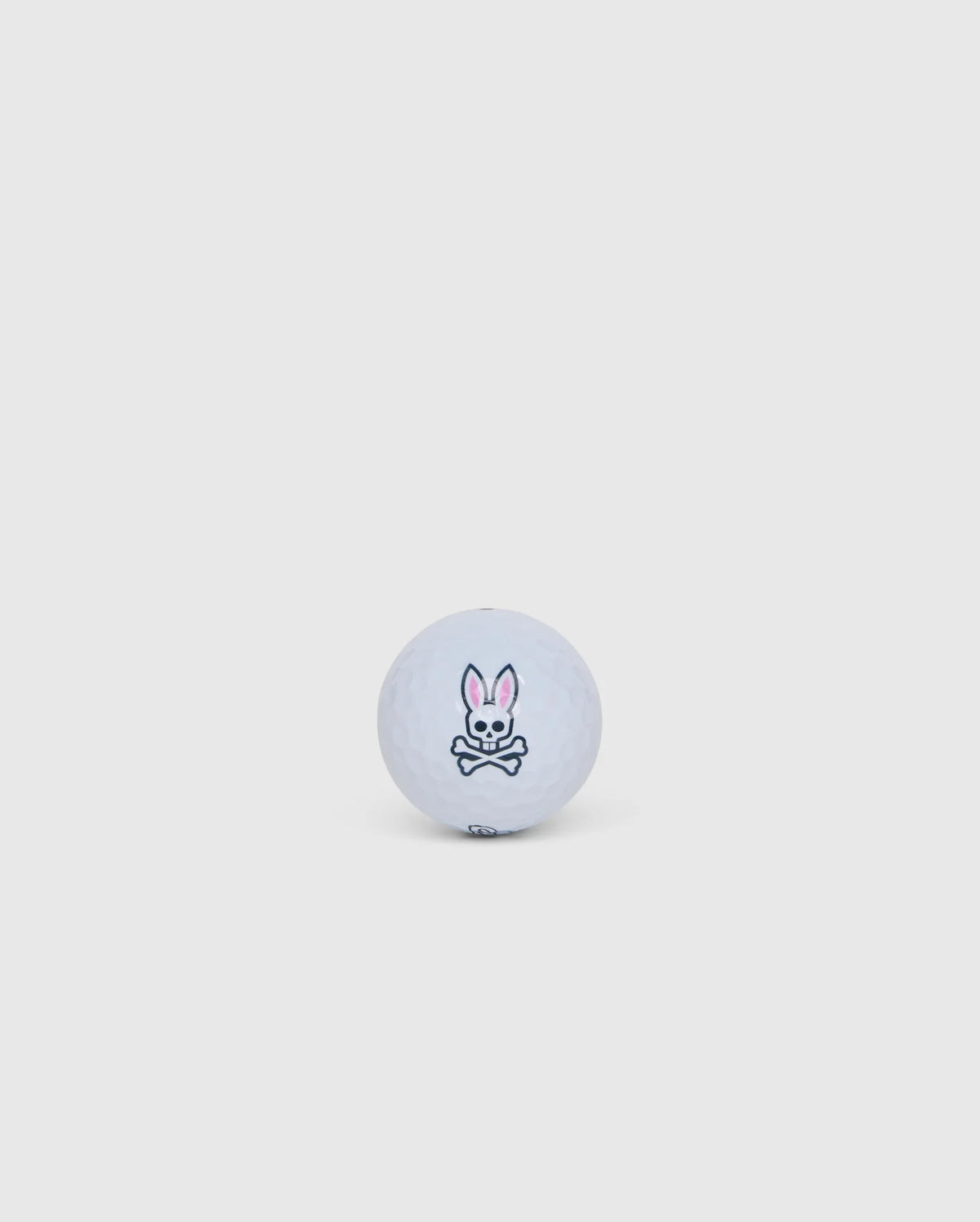 A white golf ball from the Psycho Bunny collection, featuring a simple logo with a cartoon bunny and crossed bones underneath, centered on a plain light gray background. Offered in an elegant golf ball sleeve, the 3 PACK GOLF BALLS - B6A565C200 is designed to minimize driver spin for enhanced performance.