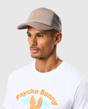 A man wearing the Psycho Bunny MENS PARIS TRUCKER CAP - B6A591C200 in beige, featuring mesh paneling, an orange Bunny logo on the front and "Psycho Bunny" on the side. He is also sporting a white t-shirt adorned with "Psycho Bunny" text and a graphic of a bunny with palm trees in the background. The backdrop is light gray.