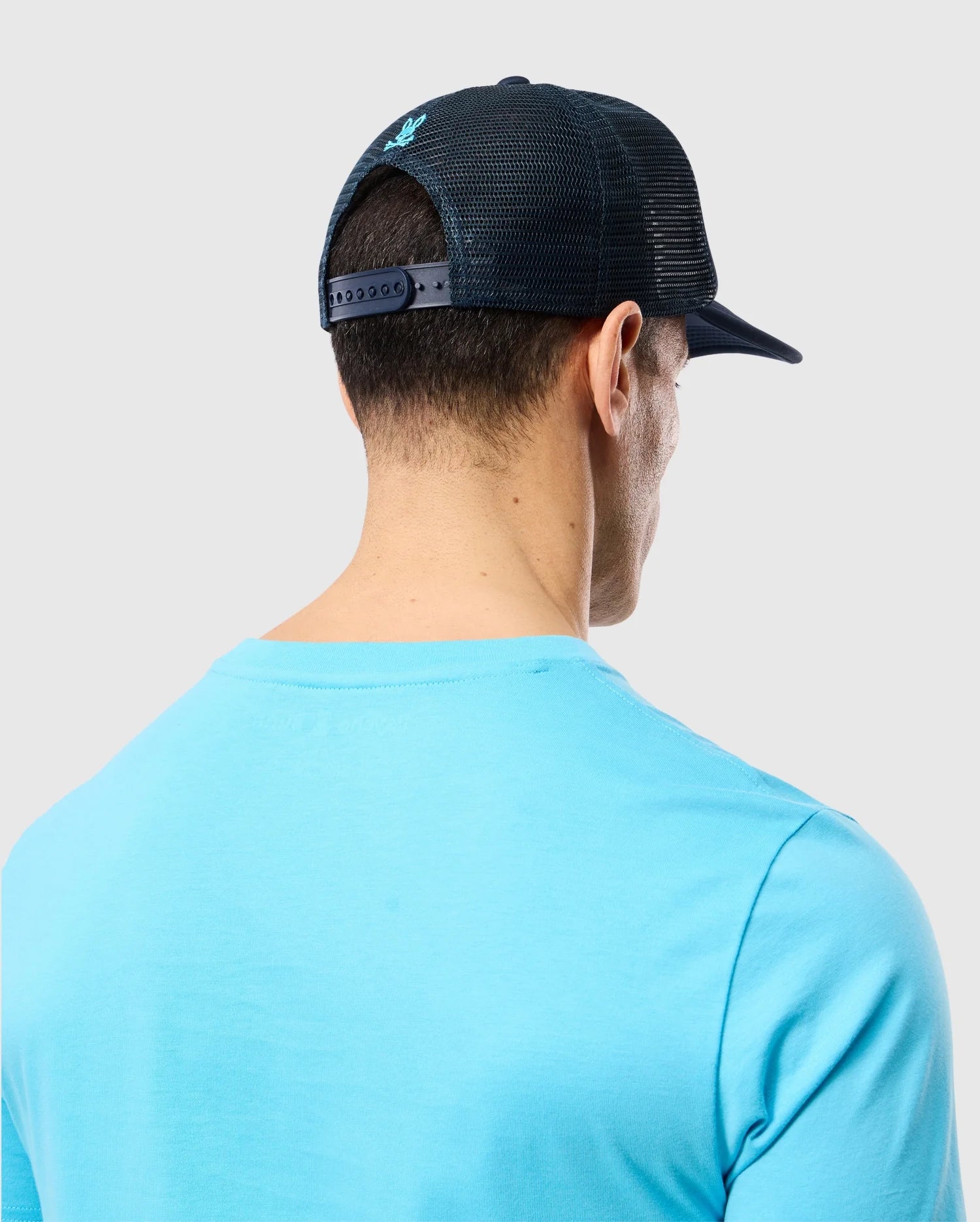 A man is seen from behind wearing a light blue T-shirt and a black trucker cap with mesh paneling. He has short dark hair visible through the mesh part of the cap, which features a subtle Bunny logo. The background is a plain light grey. The cap he is wearing is the MENS PARIS TRUCKER CAP - B6A591C200 by Psycho Bunny.