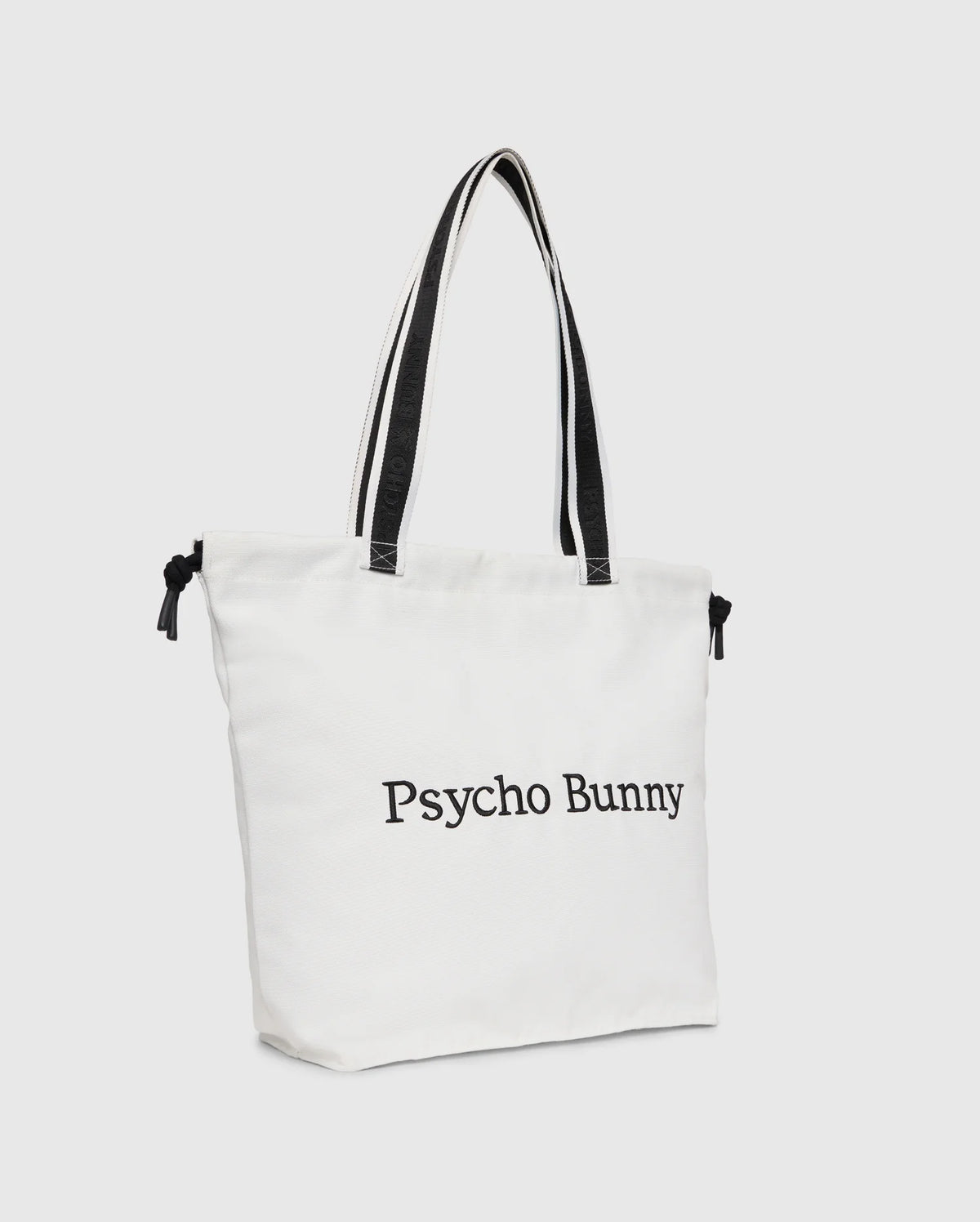 The "TENNIS TOTE - B6A596C200" by Psycho Bunny is a white canvas tote bag with black handles, featuring the text "Psycho Bunny" on the front and an embroidered logo for an elegant touch.