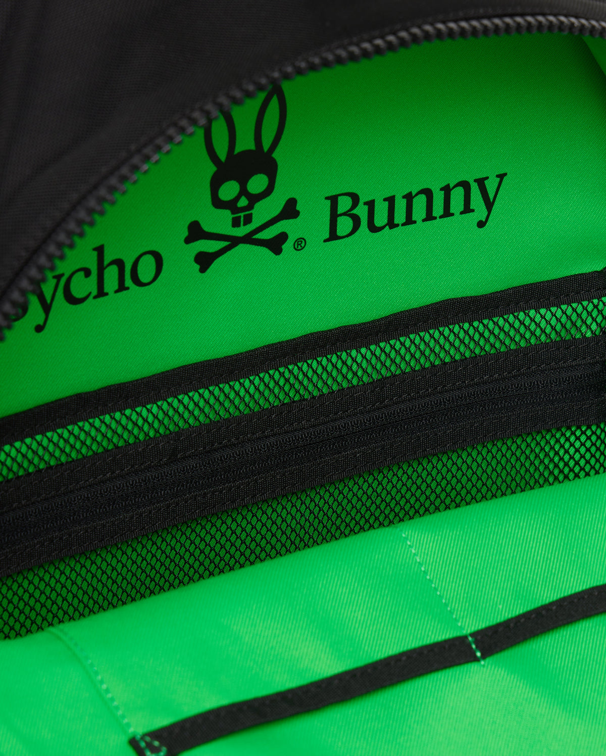 Close-up view of a vibrant green section of an Psycho Bunny Dome Backpack featuring a black graphic logo of a bunny with a skull and crossbones and the text "psycho bunny." Black zippers and mesh detail.