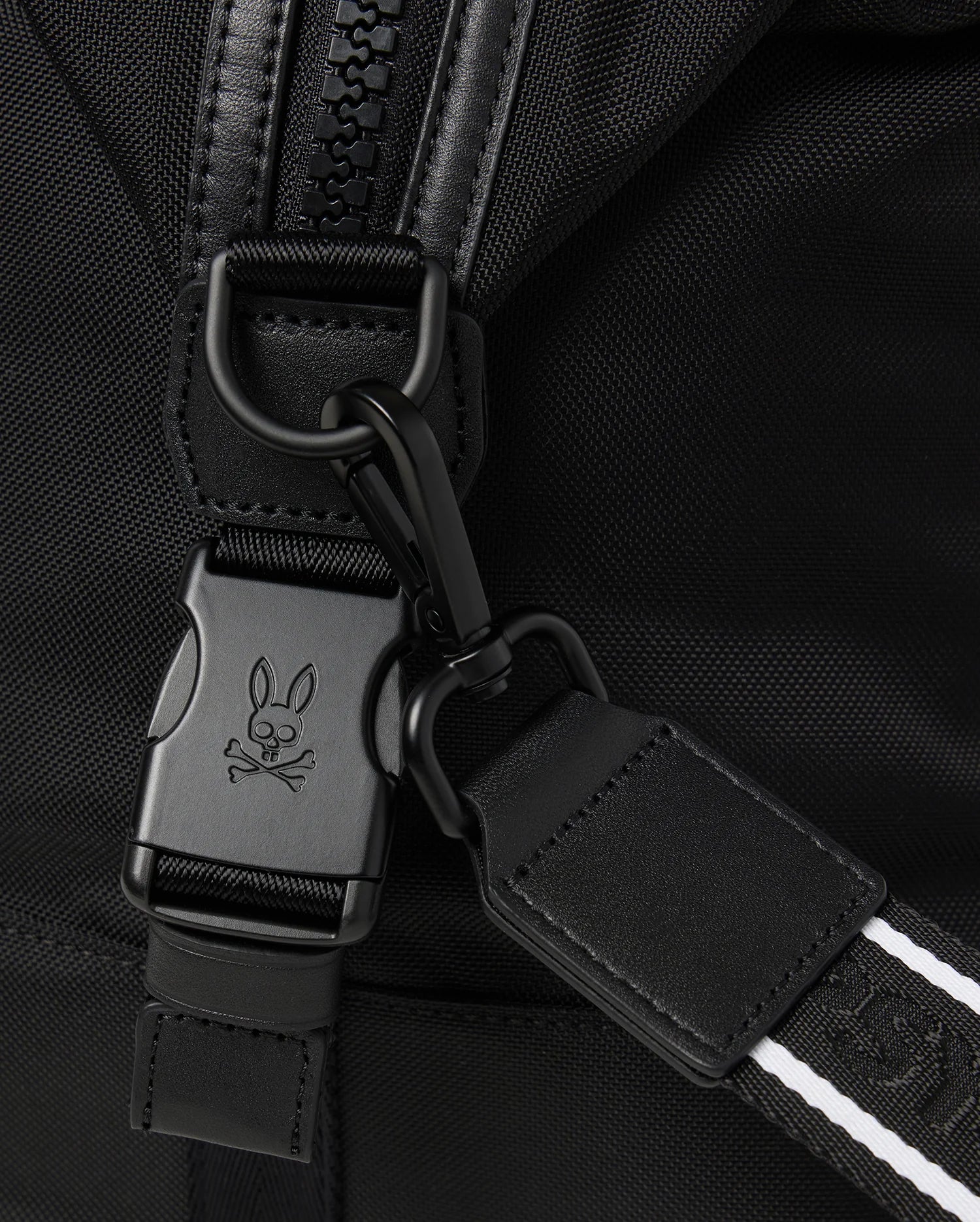 Close-up image of a backpack strap featuring a black buckle with a bunny and crossbones logo engraved on it. The strap includes a black carabiner clip and a white-striped detail to enhance its design, making it perfect for pairing with your Psycho Bunny MENS WEEKENDER BAG - B6A629Z1BG or travel luggage. The fabric is black and textured.