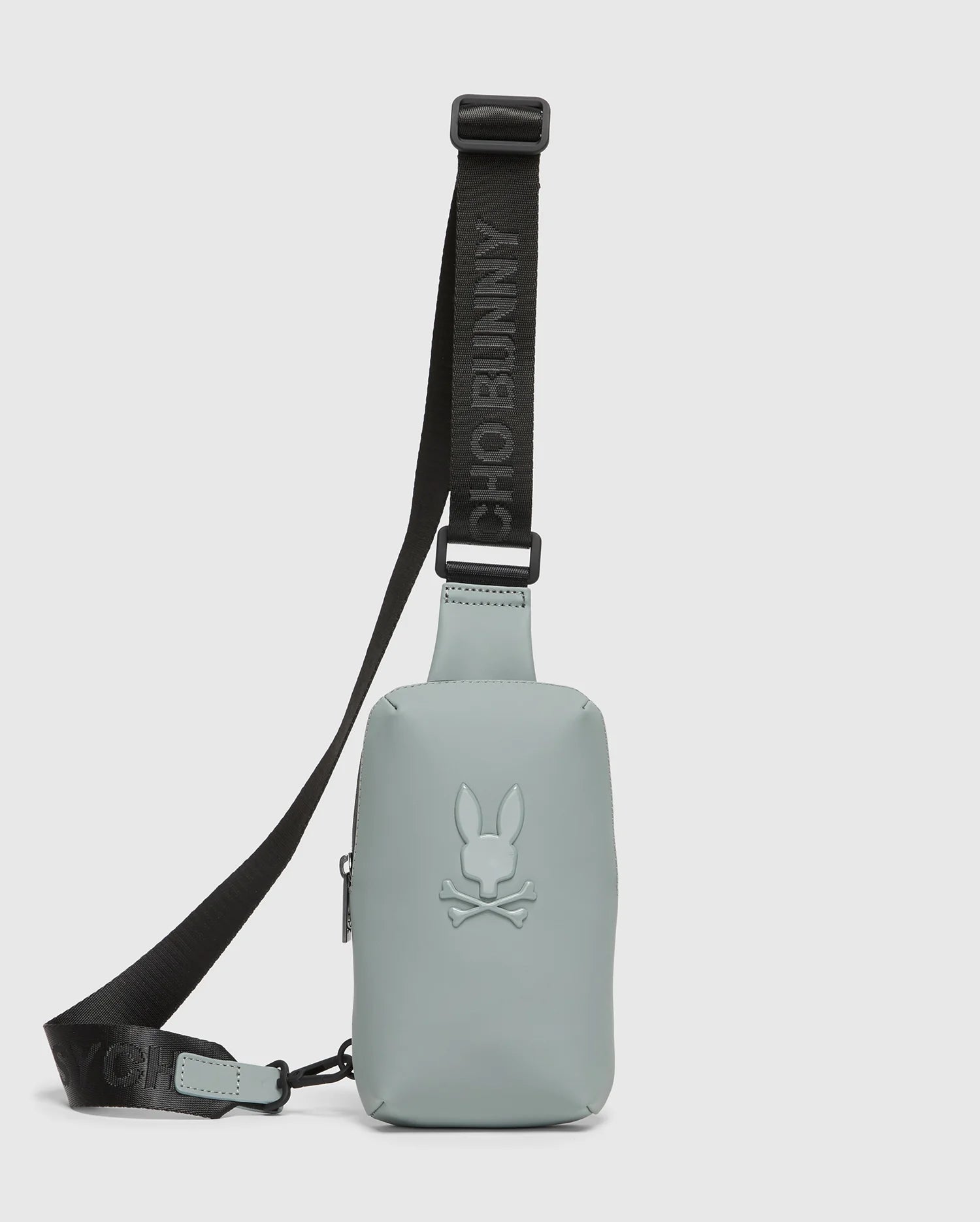 The MENS RUBBERIZED CROSSBODY - B6A635F200 by Psycho Bunny is a durable, pastel green bag made of sturdy polyester with a black adjustable strap. It features an embossed bunny with crossed bones logo and the strap boldly displays "PSYCHO BUNNY" in black letters.
